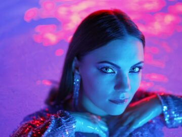 Beautiful woman posing in pool water under neon color light. Party, attractive chic in shining dress