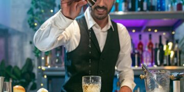 Expert barman in a white shirt and black apron smiling happy making cocktail at party in nightclub