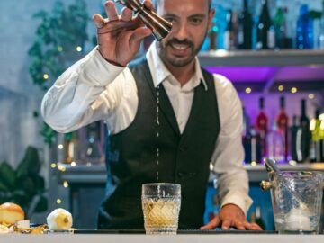 Expert barman in a white shirt and black apron smiling happy making cocktail at party in nightclub