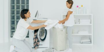 Happy mother loads clothes in washing machine, little girl helps, gives white linen from basket