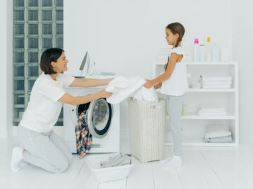 Happy mother loads clothes in washing machine, little girl helps, gives white linen from basket
