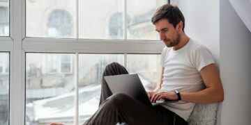 Young handsome man sits on wooden windowsill near the window in lotus position, works on gray laptop