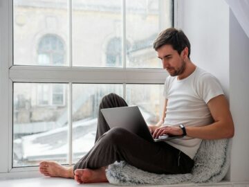 Young handsome man sits on wooden windowsill near the window in lotus position, works on gray laptop